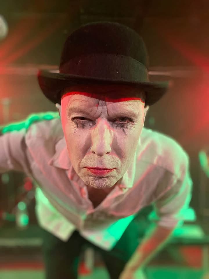 Artist med vitt målat ansikte, mörka tårstreck och svart plommonstop lutar sig mot kameran