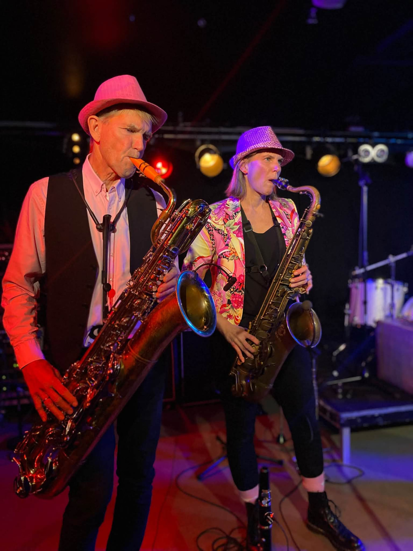 Två musiker i hattar spelar saxofon tillsammans på scen med strålkastare bakom