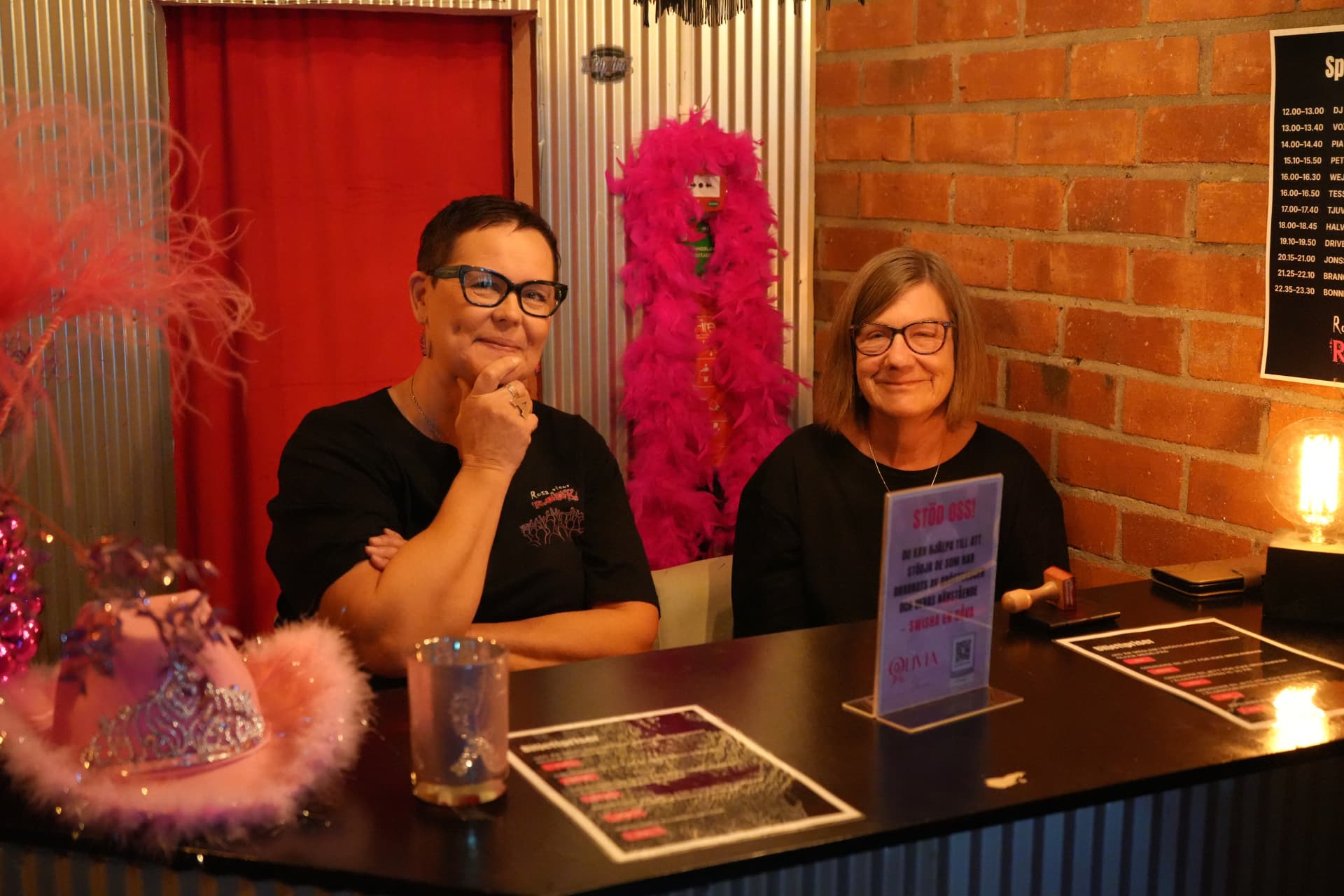 Två kvinnor bakom entrédisken med en rosa hatt med fjäderboa, informationsblad och en skylt om att swisha en gåva till Olivia
