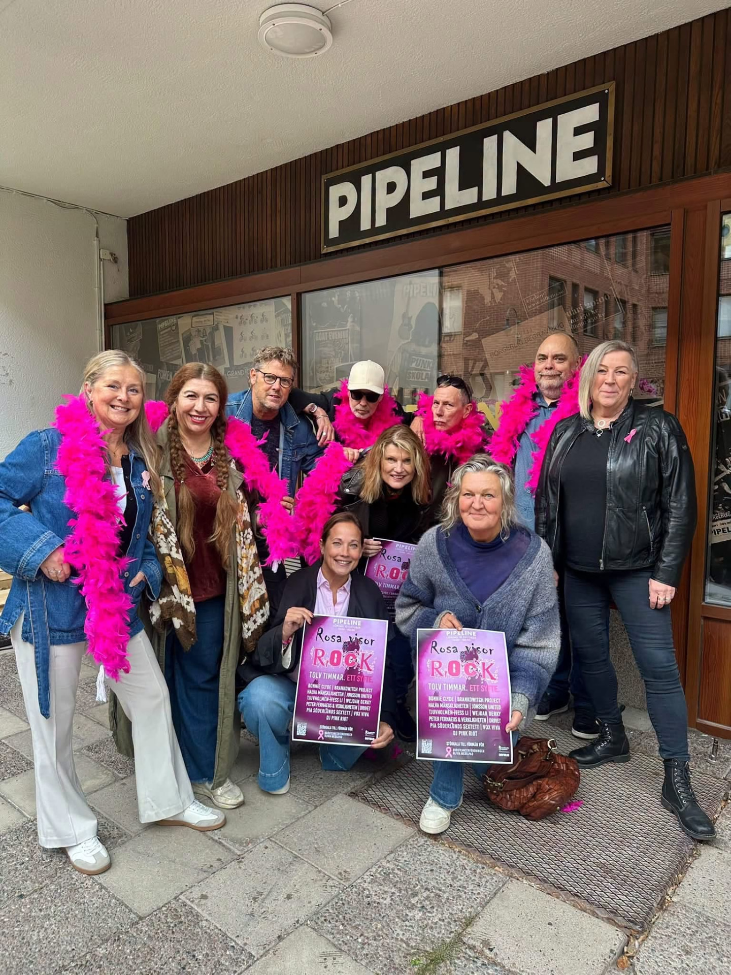 Grupp galagäster med rosa fjäderboor utanför Pipelines entré, två personer som sitter på huk håller varsin rosa affisch om galan