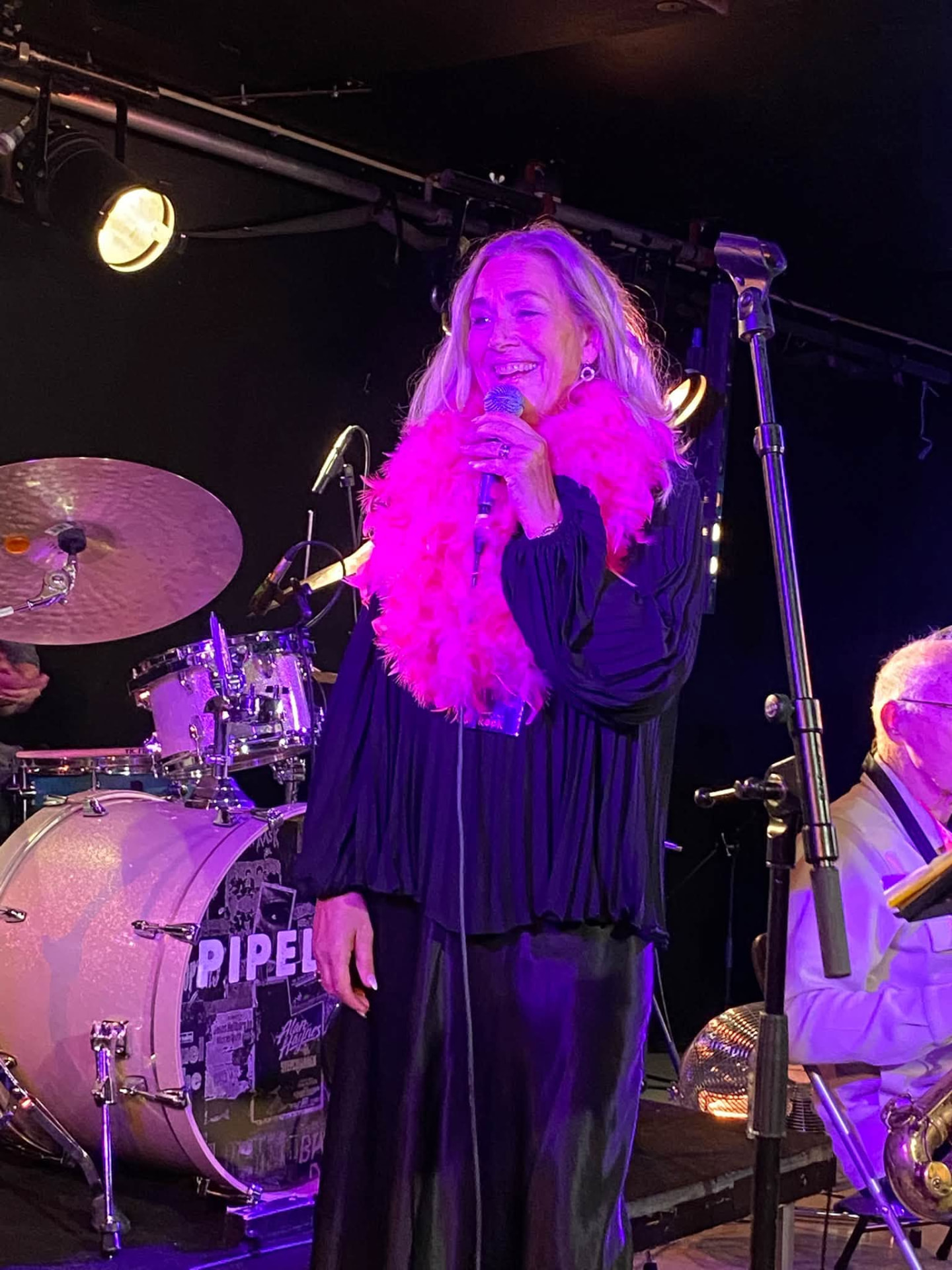 Leende sångerska med mikrofon och ceriserosa fjäderboa på scen framför ett trumset, bredvid en musiker i lila och varma scenljus
