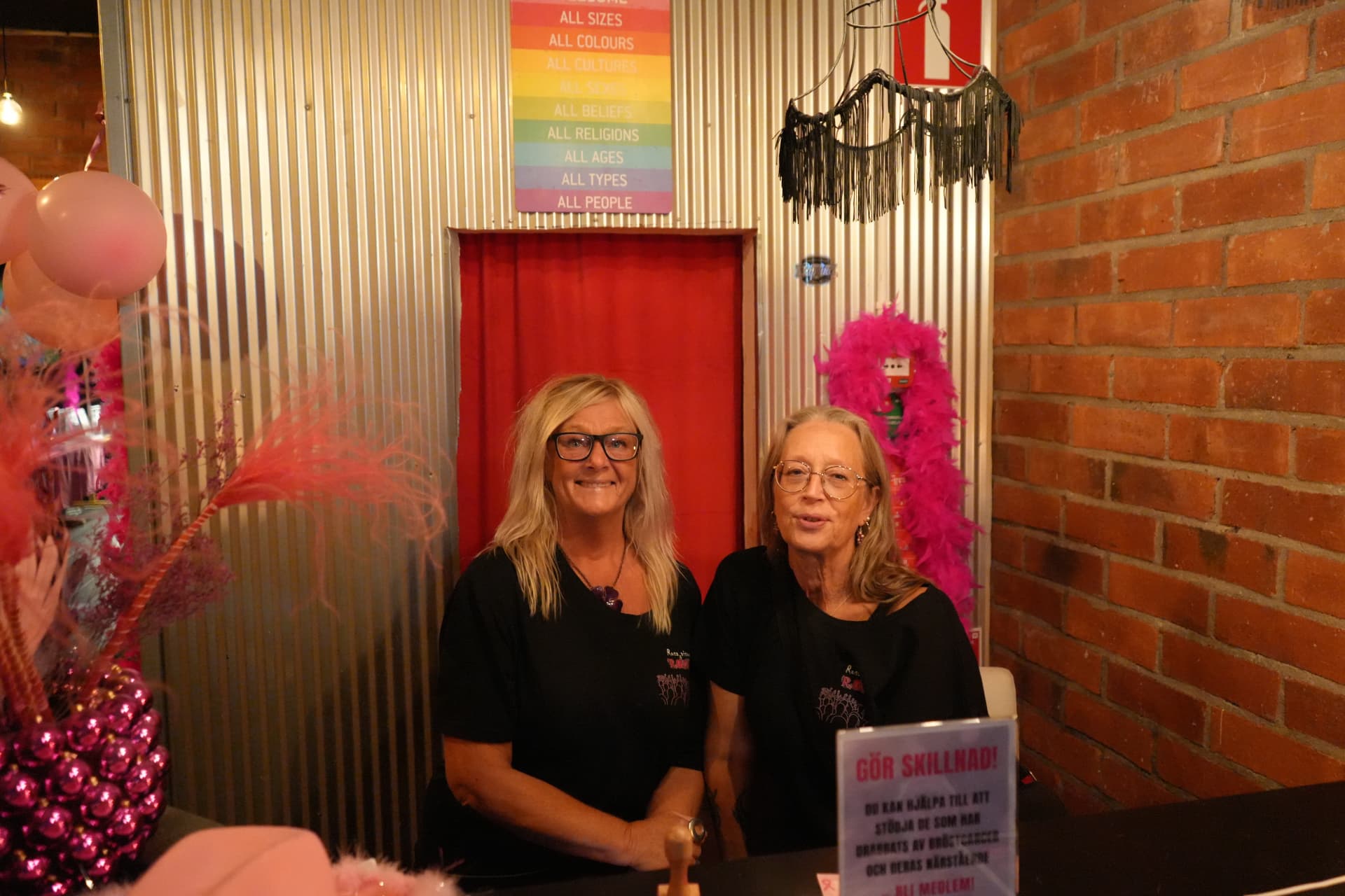 Två kvinnor bakom entrédisken bland rosa ballonger, fjäderboor och en informationsskylt