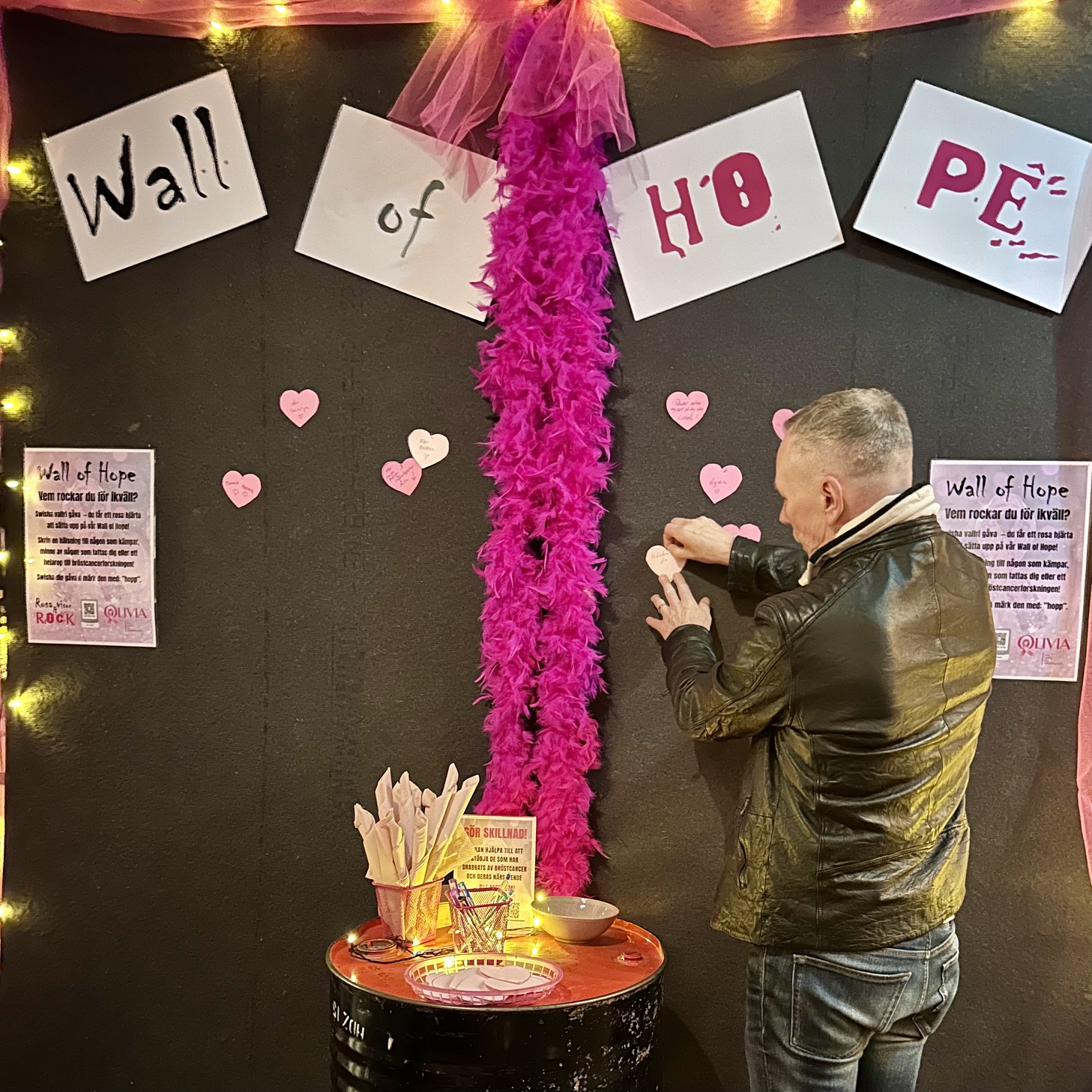 Man i svart läderjacka sätter upp ett rosa pappershjärta på Wall of Hope-väggen, med rosa fjäderboa, skyltar och ljusslingor