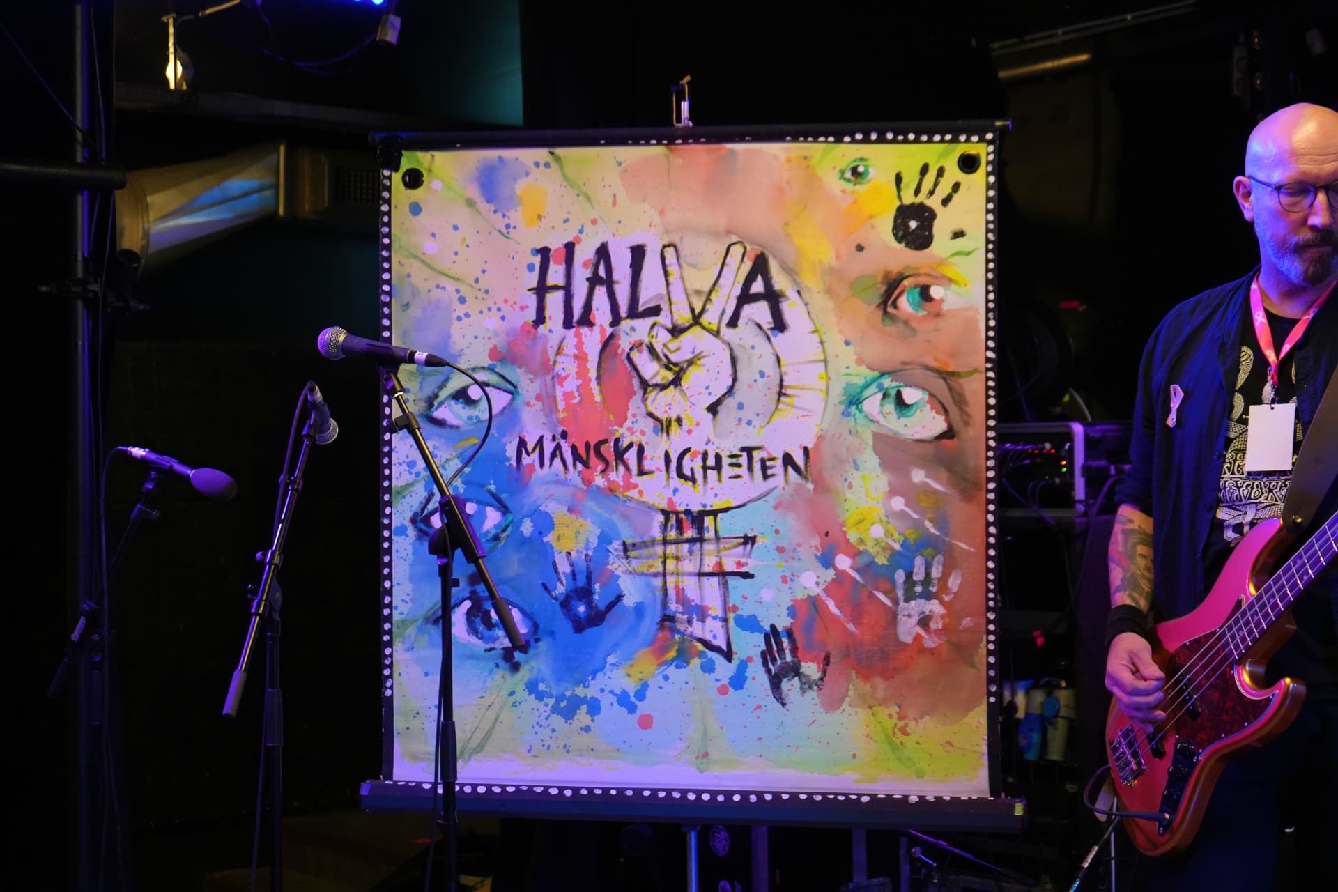 Musiker med glasögon och skägg spelar röd elbas på scen framför en målad duk med texten Halva Mänskligheten