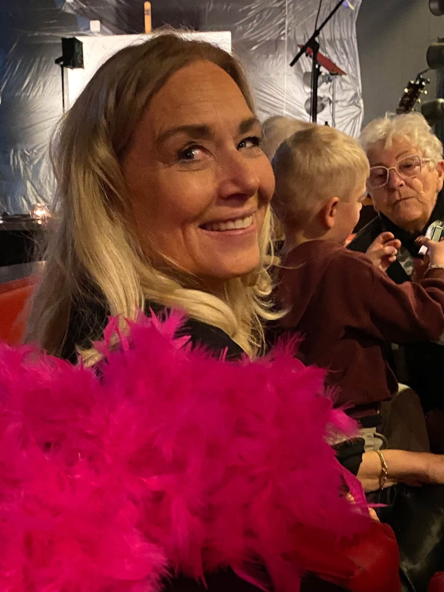 Leende galagäst sedd från sidan med ceriserosa fjäderboor i förgrunden, barn och annan publik bakom