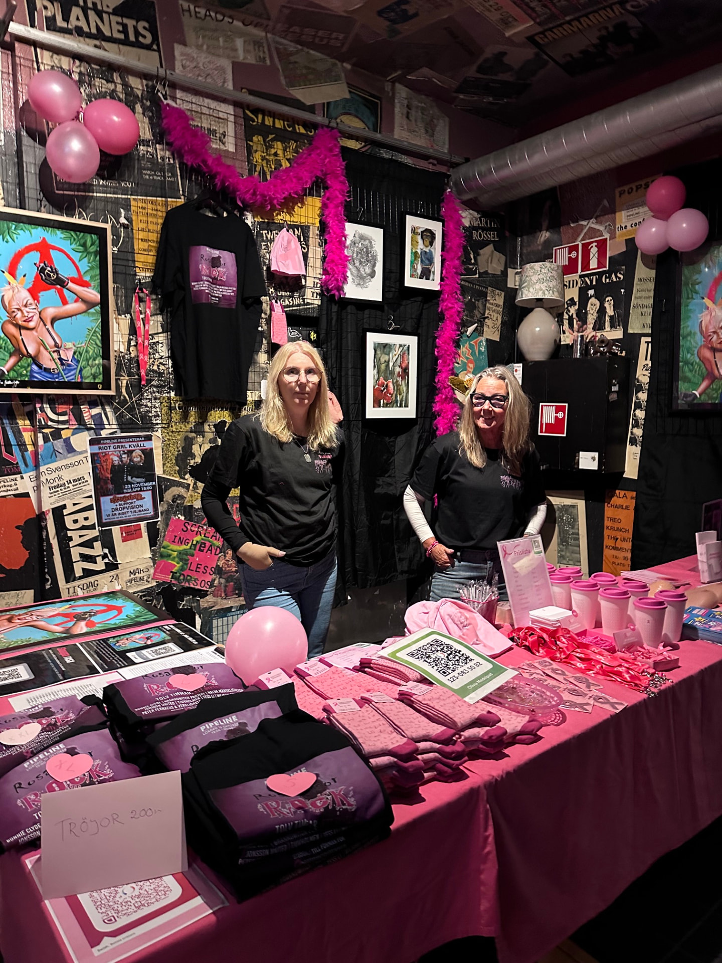 Två kvinnor i svarta t-shirt bakom ett bord med rosa duk, galatröjor, rosa sockor, mössor och Rosa bandet-produkter