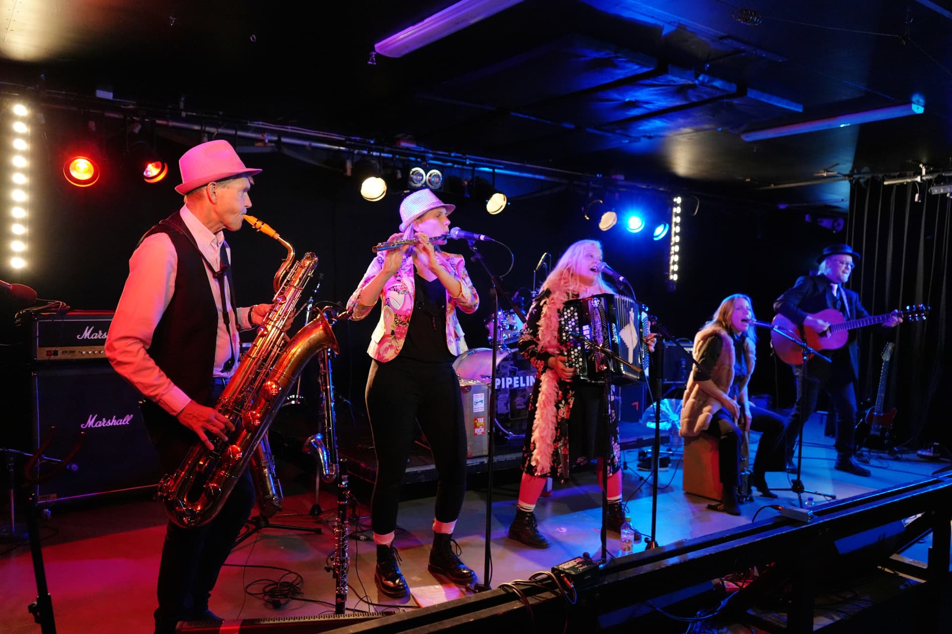 Fem musiker spelar på scen: saxofonist i rosa hatt, tvärflöjtist, dragspelerska med rosa fjäderboa, sångerska som sitter på en cajon och gitarrist i mörk kostym