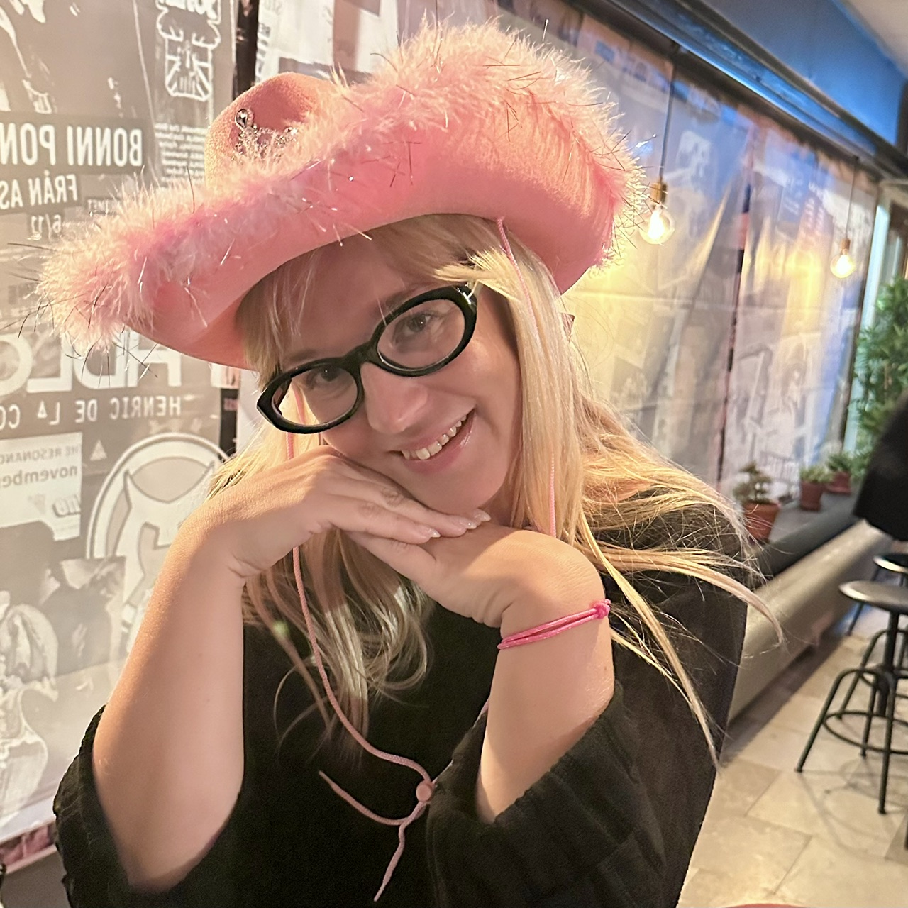 Leende kvinna med svartbågade glasögon och rosa cowboyhatt med fjäderbrätte och tiara