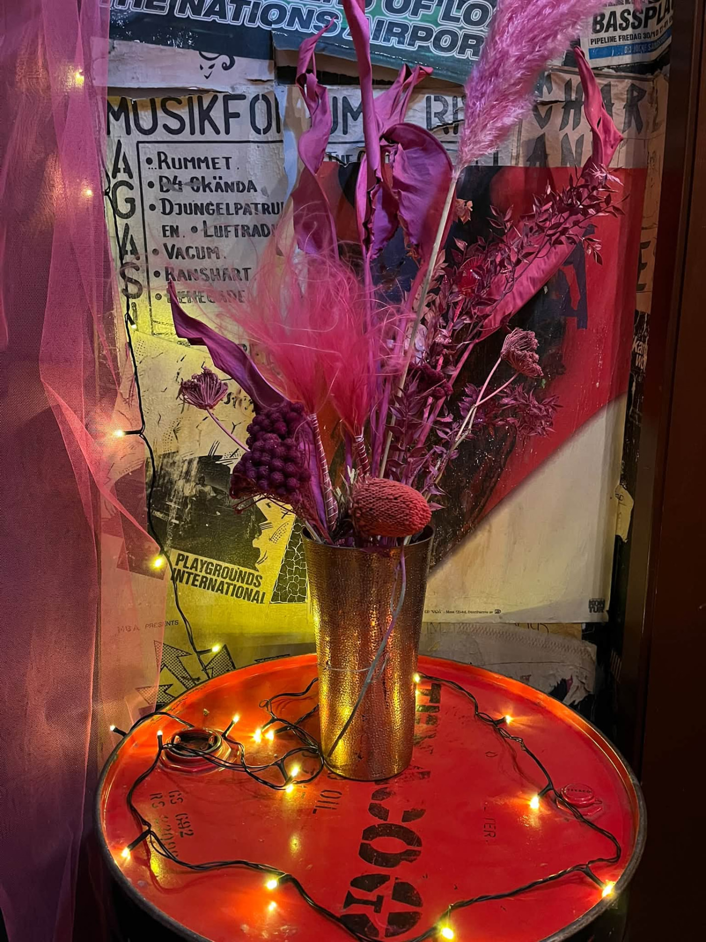 Guldfärgad vas med rosa fjädrar och torkade växter på ett rött plåtfat omgiven av en tänd ljusslinga, framför en affischtäckt vägg och rosa tyll