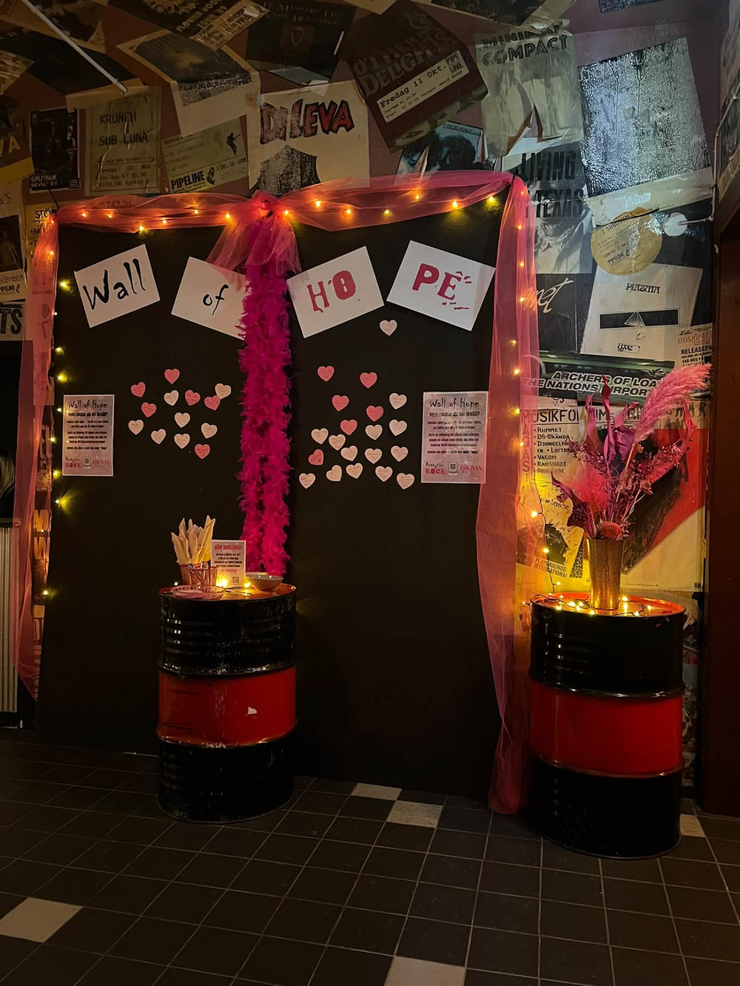 Vägginstallation med texten Wall of HOPE i vita papper, rosa pappershjärtan och en ceriserosa fjäderboa, inramad av rosa tyll, ljusslingor och två plåttunnor med vas och lappar
