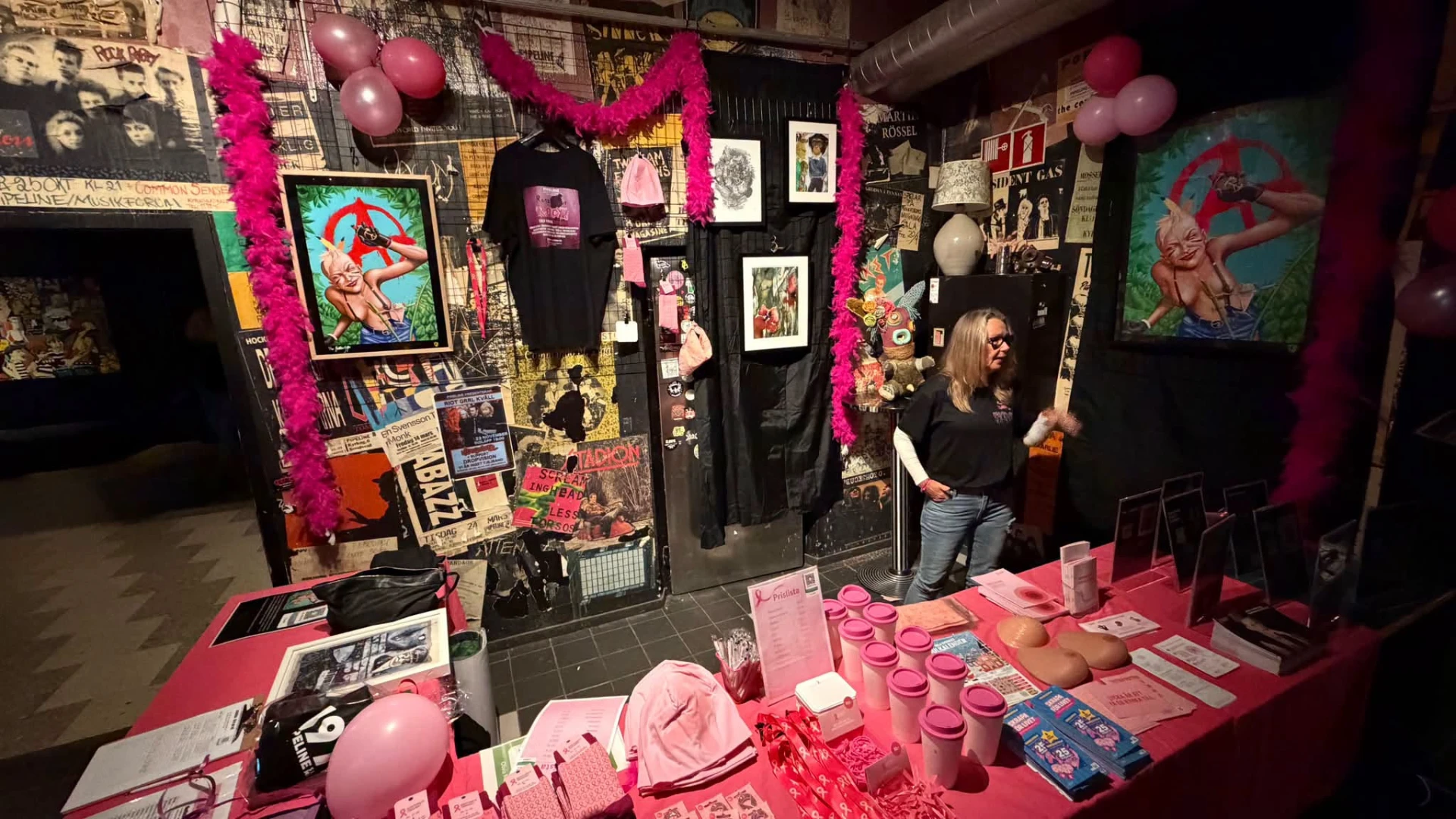 Kvinna bakom ett informations- och försäljningsbord i rosa tema med muggar, mössor och ballonger
