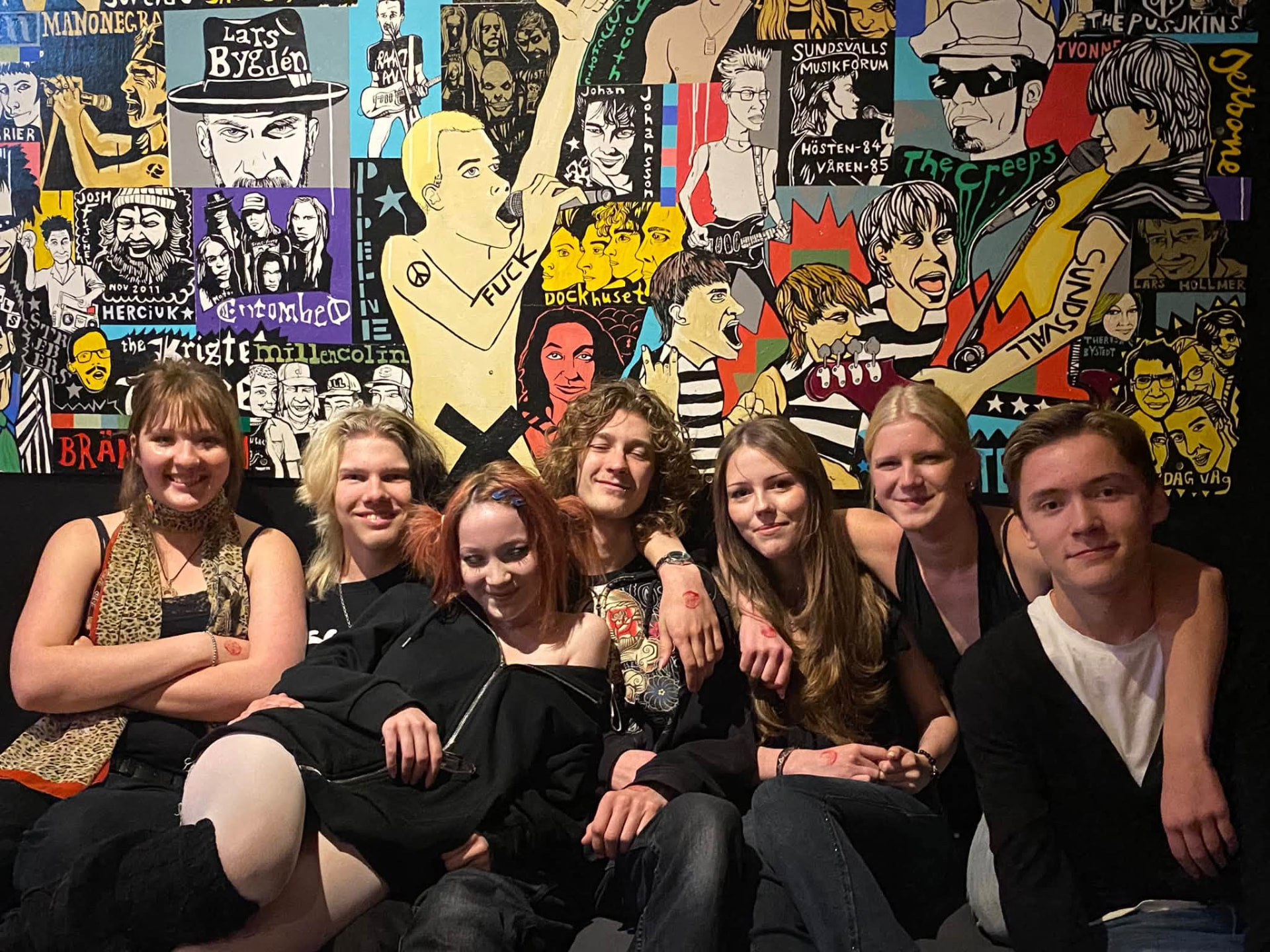 Grupp ungdomar som sitter i en soffa framför en illustrerad musikvägg med bandnamn