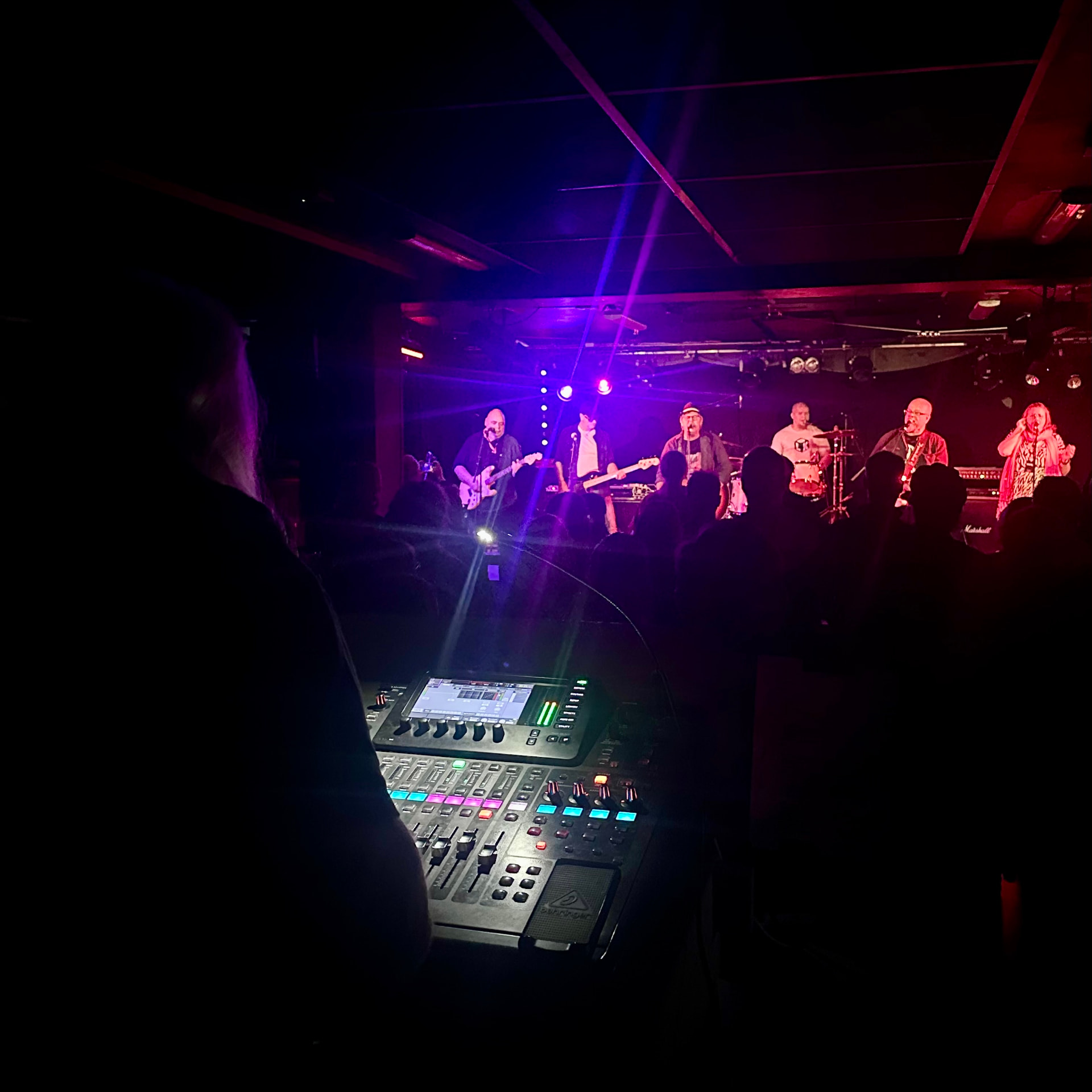 Band på scen i rött och lila ljus sett från mixerpulten, publik i förgrunden