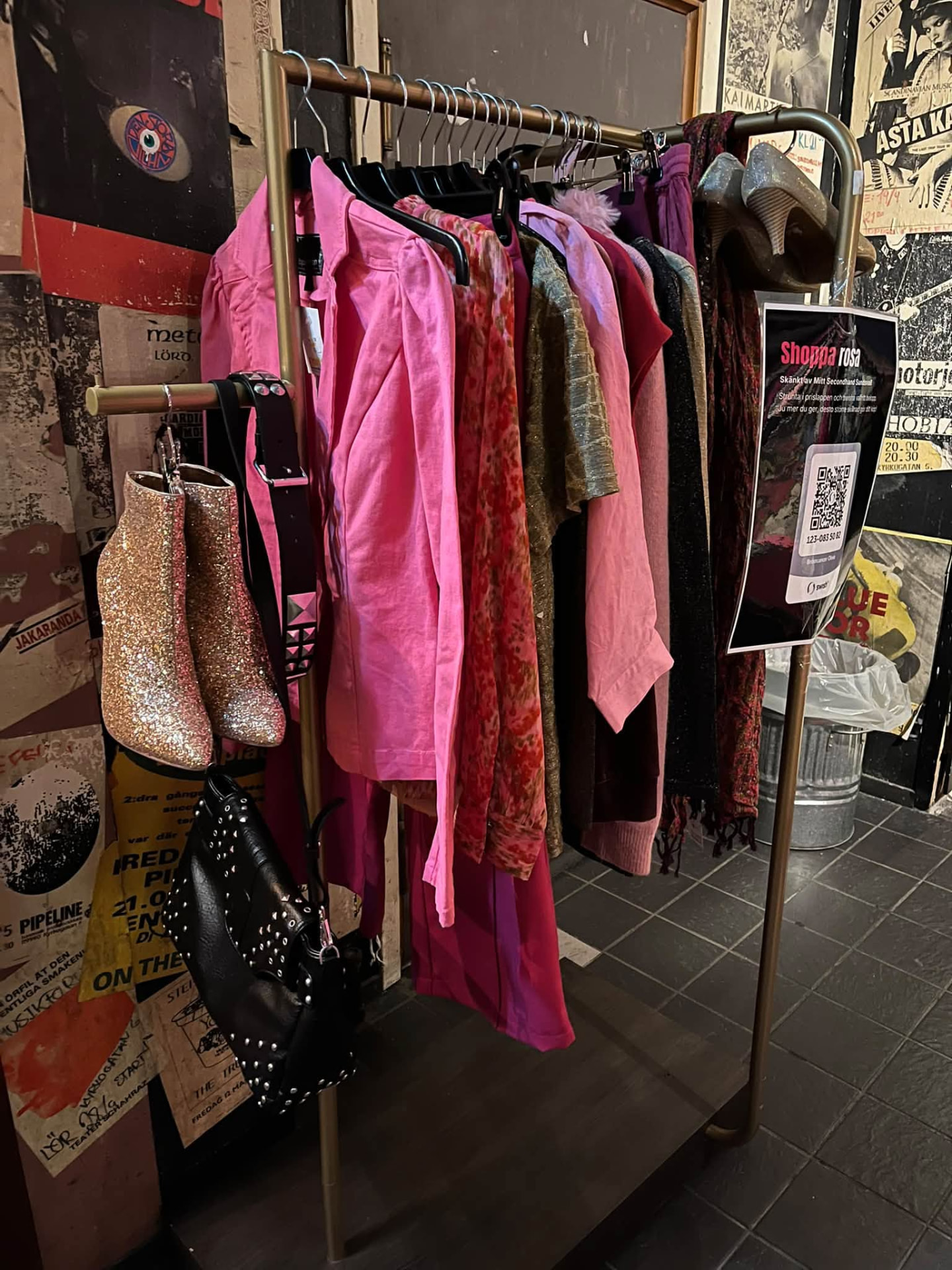 Klädställning med rosa och mörka plagg, hängande guldglittriga stövletter, svart nitad handväska och en skylt med texten Shoppa rosa