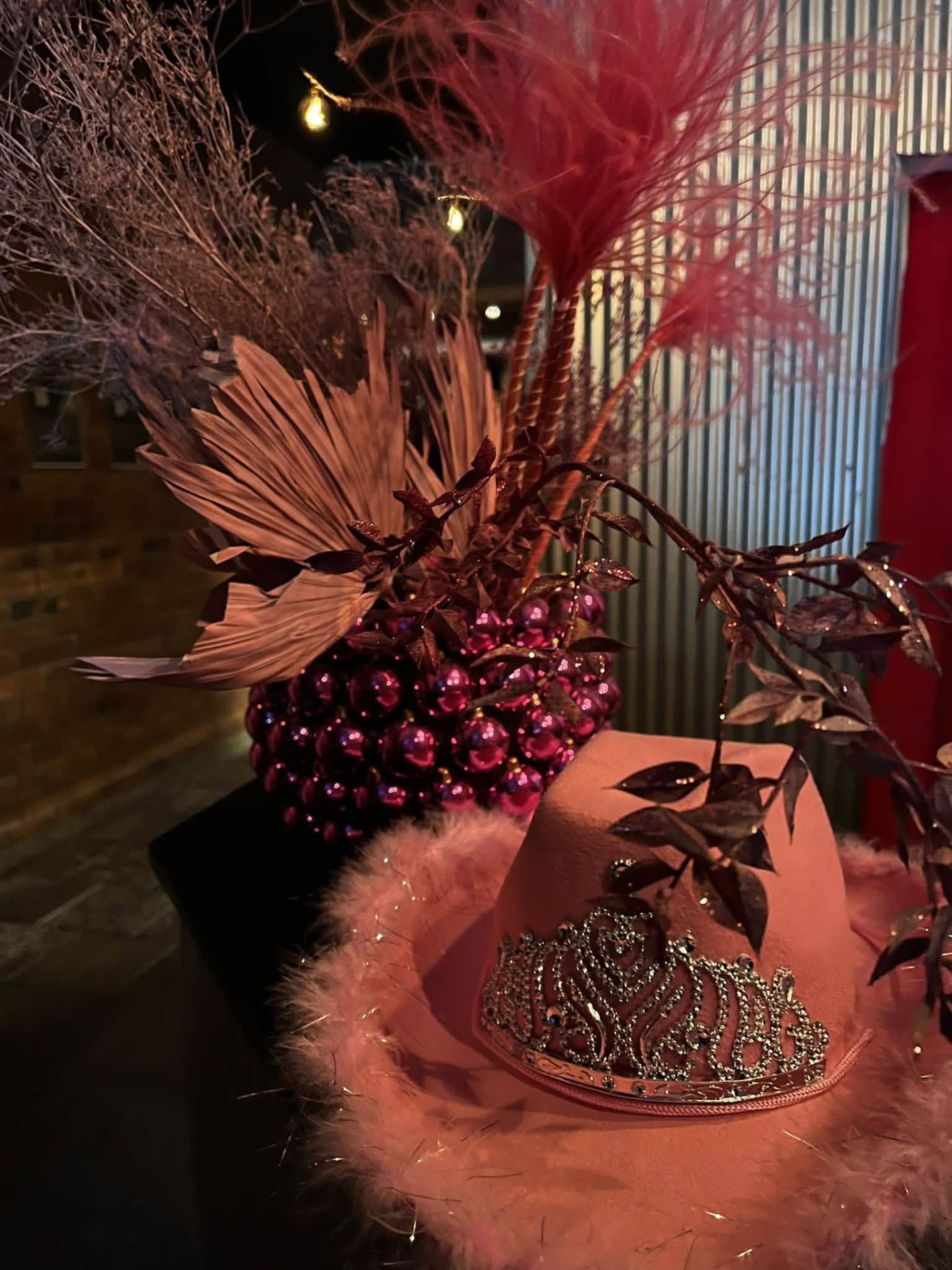 Rosa cowboyhatt med fjäderbrätte och strasstiara framför ett arrangemang av ceriserosa julgranskulor och torkade växter